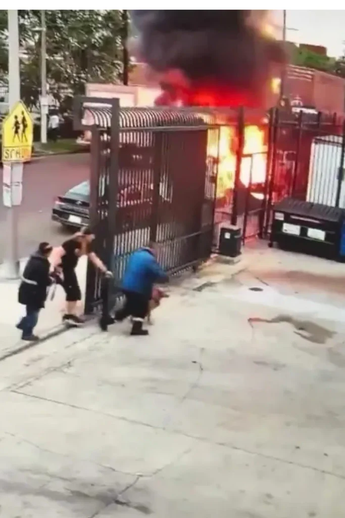 Man carrying a dog to safety as a van burns behind a gate on an LA street