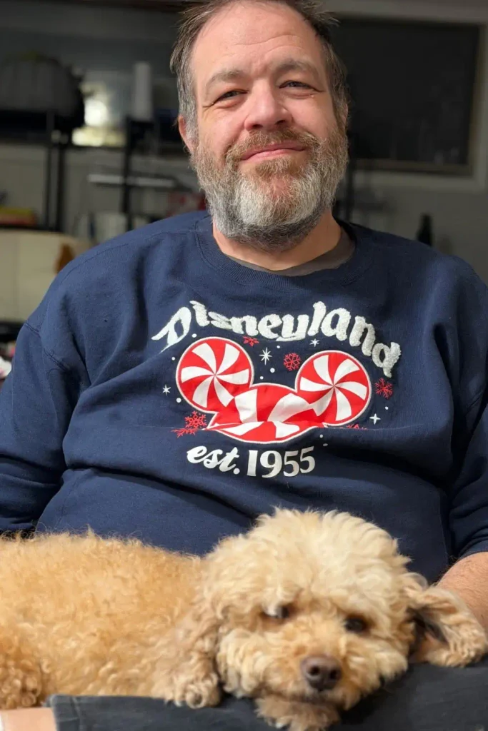 Smiling bearded man in a navy sweatshirt with a curly tan dog resting its head on his lap